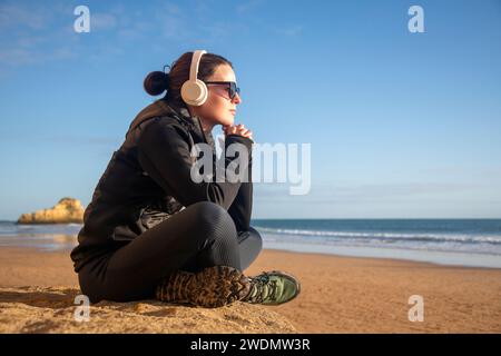 woman sitting on a rock looking out to sea, listening to music on headphones, winter sun. Stock Photo