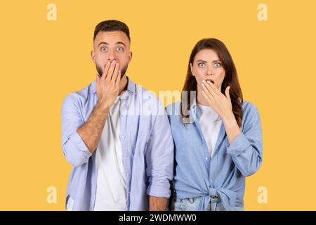 Shocked couple covering mouths, wide-eyed surprise on yellow background Stock Photo