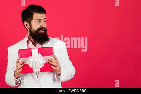 Bearded man in white suit with present box look aside on copy space for advertising. Birthday or anniversary. Smiling businessman in formal wear with Stock Photo