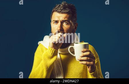 Ill bearded man with flu and fever drinking medicine tea. Handsome guy having sore throat. Sick man in sweater wrapped in scarf with cup hot healing Stock Photo