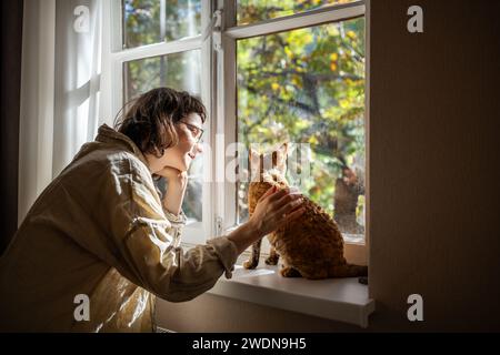 Tender pet lover stroking beloved adorable breed cat Devon Rex sitting on windowsill in sunlight Stock Photo