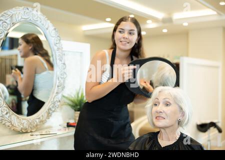 Young woman hairdresser holds mirror behind senior female client and shows result of work. Stock Photo