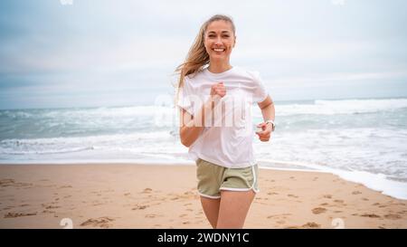 Smiling female athlete jogging on sandy sea beach. Fit woman in white top and sporty shorts. Active lady running along shoreline represents healthy lifestyle. Stock Photo