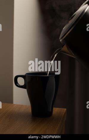 Pour hot water from an electric kettle into a dark blue ceramic cup on a wooden table in the middle of the night. Stock Photo