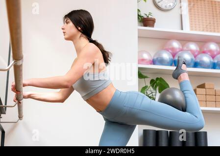 Side view of confident young female athlete in sportswear looking away while standing with hands on barre and doing exercises with fit ball in gym Stock Photo