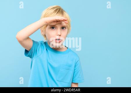 Cute preschooler looking curiously into distance with hand on the forehead. Kid looking into future, studio portrait on pastel blue background. Stock Photo