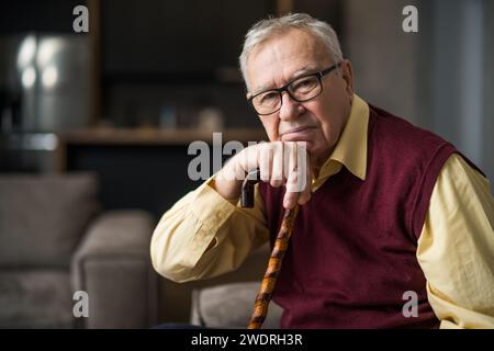 Pensive and worried senior man is sitting on bed in his home. Portrait of senior man with walking cane. Stock Photo