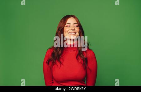 Portrait of a young, stylish female with a colorful sweater and nose ring stands confidently against a vibrant green background. Her cheerful smile ex Stock Photo