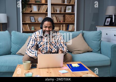 Young African American man with headset having web call using laptop computer and smartphone sitting on sofa talking at webcam in home office Video co Stock Photo