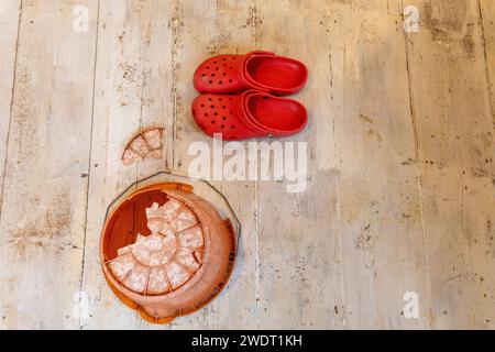 On whitewashed floorboards an inverted orange plastic bucket with an enormous hole in the bottom and a pair of bright red Crocs Stock Photo