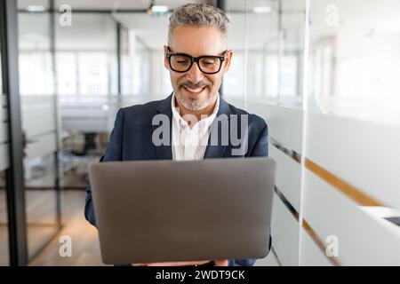 Smart and intelligent senior 60s businessman wearing formal suit and white shirt using laptop indoors, male office employee holding computer and typing message, smiling, enjoying his job Stock Photo