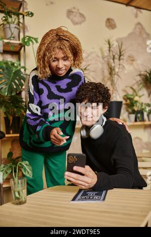 happy multicultural couple with curly hair looking at smartphone and choosing menu in vegan cafe Stock Photo