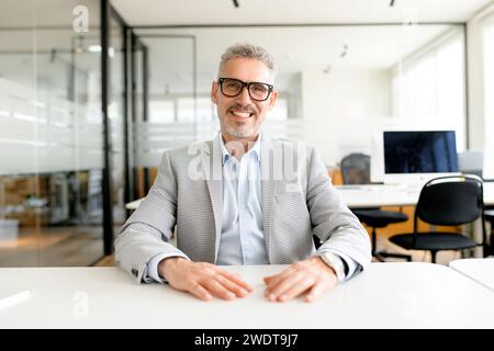 Mature businessman in a grey checkered blazer and crisp blue shirt exudes confidence as he sits at a polished office table, looking at the camera with charismatic smile Stock Photo