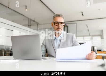 An attentive businessman reviews documents while working on his laptop in a well-lit office space, portraying the meticulous nature of business planning and analysis Stock Photo