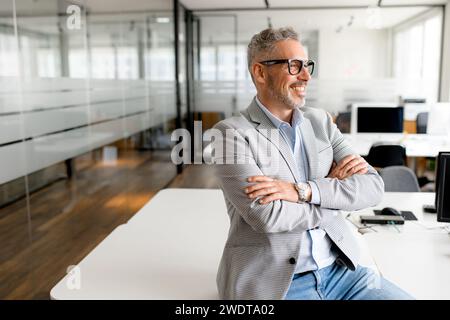 A mature businessman wearing stylish glasses and a modern grey suit leans comfortably in his desk, exuding a sense of contentment and achievement Stock Photo