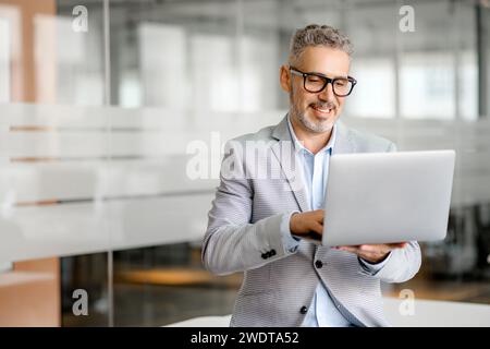 A charismatic businessman in a modern office space confidently uses a laptop, symbolizing the integration of technology in contemporary business practices. His focused demeanor showcases dedication Stock Photo