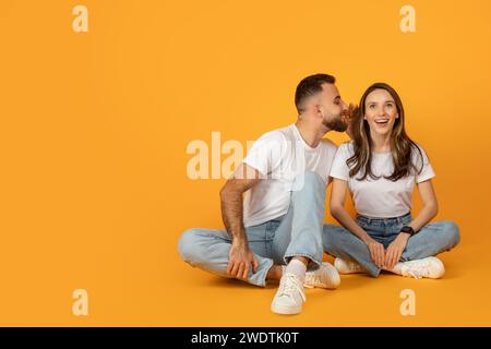 A joyful young couple in casual clothing sits cross-legged on a vibrant orange background Stock Photo