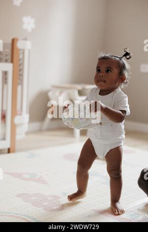 Mixed race toddler girl carrying diaper in her hands in nursery. Afro-Caucasian little girl. Concept of interracial family. Stock Photo