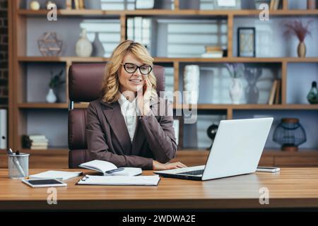 Elegant senior businesswoman with glasses smiling while working on a laptop in a modern, well-decorated home office environment. Stock Photo