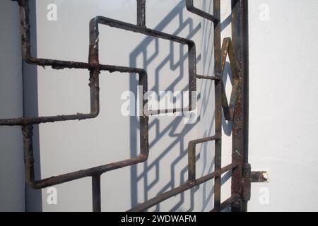 Rustic window iron grill against white wall, shadow against the white background to see the design. Paint coat is peeling off showing rust. Singapore. Stock Photo