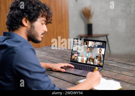 A focused Indian man sits at a wooden desk engaging in a video conference on his laptop, pen in hand, ready to jot down important points, showcasing the interactive nature of remote communication Stock Photo