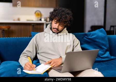 Young male Indian student or freelancer man jotting down notes from the laptop, working or studying at a distance. The comfortable home setting contrasts with the focused work activity Stock Photo