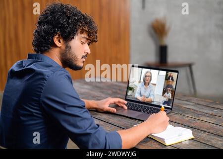 Focused young man with curly hair is participating in virtual meeting, taking notes as listens to a woman on his laptop screen. Modern remote work environment and the concept of digital communication Stock Photo