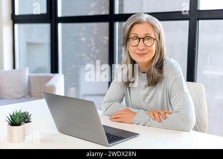 Confident mature female manager ceo sitting at desk indoor, using laptop, gray-haired senior businesswoman looking at the camera sits in bright modern office Stock Photo