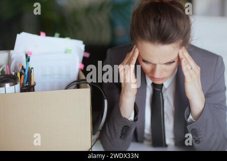 New job. pensive modern middle aged woman worker in modern green office in grey business suit with personal belongings in cardboard box. Stock Photo