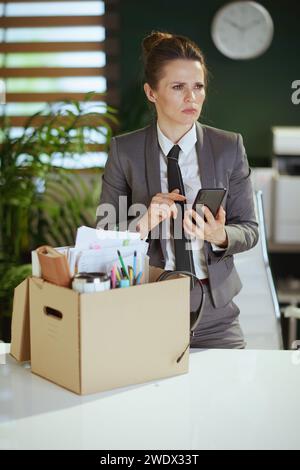 New job. pensive modern 40 years old woman worker in modern green office in grey business suit with personal belongings in cardboard box talking on a Stock Photo