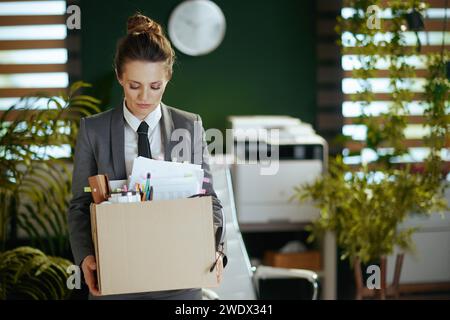 New job. sad modern woman worker in modern green office in grey business suit with personal belongings in cardboard box. Stock Photo