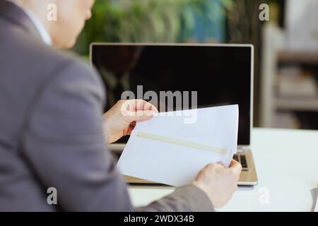 New job. Seen from behind modern middle aged woman worker in modern green office with laptop opening letter. Stock Photo