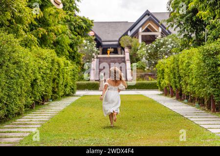 Young girl in white dress running towards house along garden path. Childhood and freedom. Stock Photo
