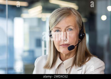 Close-up portrait of an old gray-haired woman wearing a headset sitting in the office, looking seriously and confidently at the camera. Stock Photo