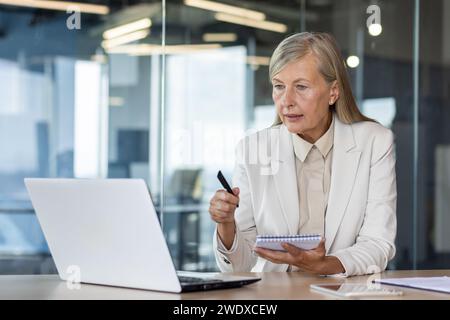 Serious senior businesswoman, executive director works in the office at the laptop, sits at the table, communicates via video call, makes notes in a notebook. Stock Photo