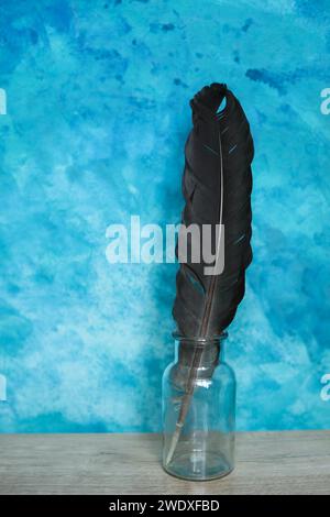 Black feather in a glass bottle on a wooden table against a blue background Stock Photo