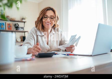 Beautiful middle-aged woman in glasses and paper bills joyfully laughing while calculating revenue grows at home office. Small business, successful in Stock Photo