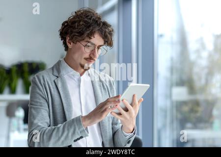 Businessman young thinking serious and concentrated standing near the window inside the office, the man is holding a tablet computer in hands, using application, passionately reading from screen. Stock Photo