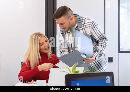 A smiling woman and a man holding a folder review a document together in a light-filled office space. Stock Photo
