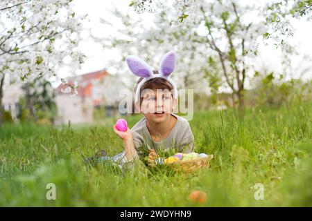 Easter egg hunt. Cute smiling boy in bunny ears lying on green grass with basket of Easter eggs at Easter egg hunt in garden. Portrait against backgro Stock Photo