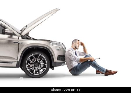Unhappy driver sitting on the ground in front of a SUV with open hood isolated on white background Stock Photo