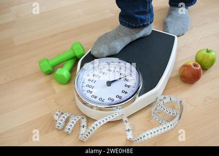 Feet of a man in gray socks stepping on an analog weight scale, green dumbbells, measuring tape and apples lying nearby on a wooden floor, fitness and Stock Photo