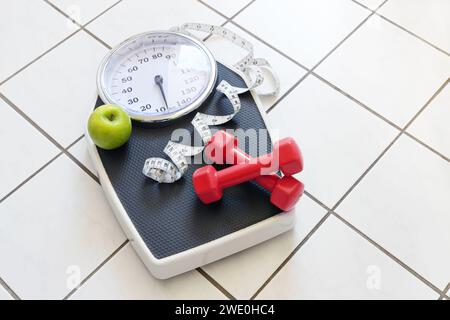 Personal scale with red dumbbells, measuring tape and a green apple on a tiled floor, fitness and weight loss concept, copy space, selected focus Stock Photo