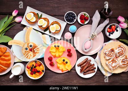 Fun Easter breakfast or brunch table scene. Overhead view on a dark wood background. Bunny pancake, egg nests, chick fruit and a variety of spring foo Stock Photo