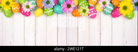 Colorful Easter eggs and spring flowers. Overhead view top border against a white wood banner background. Copy space. Stock Photo