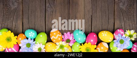 Colorful Easter eggs and spring flowers. Top view bottom border against a dark wood banner background. Copy space. Stock Photo