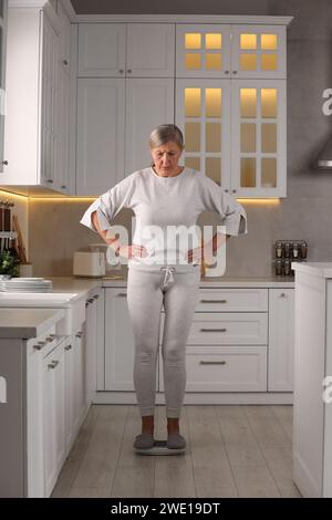 Menopause, weight gain. Concerned woman standing on floor scales in kitchen Stock Photo