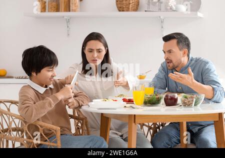 Teenage boy using tablet computer and his angry parents at table during dinner in kitchen. Family problem concept Stock Photo