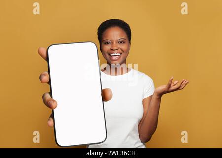 Joyful woman showing smartphone with empty blank screen display. Model in white t-shirt with phone on yellow studio wall background Stock Photo