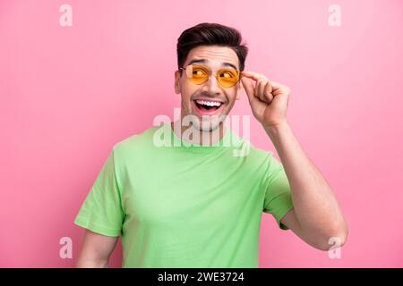 Photo of young macho smiling guy trying new sunglasses oakley wearing green t shirt looks empty space isolated on pink color background Stock Photo